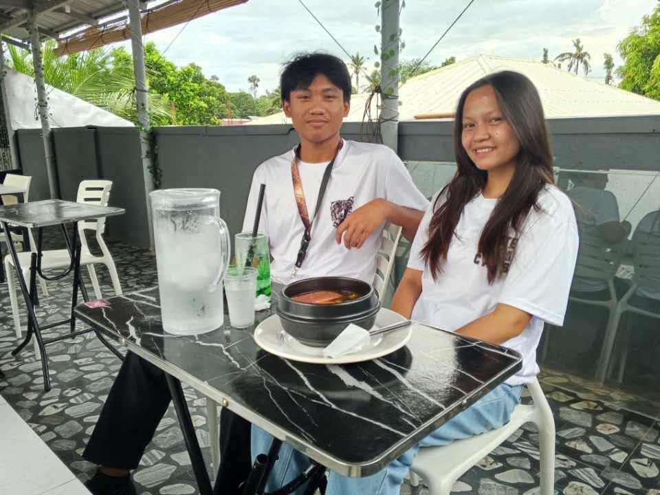 A person and a teenager sitting at a cafe table with a black bowl of food and a pitcher of water in an outdoor setting.
