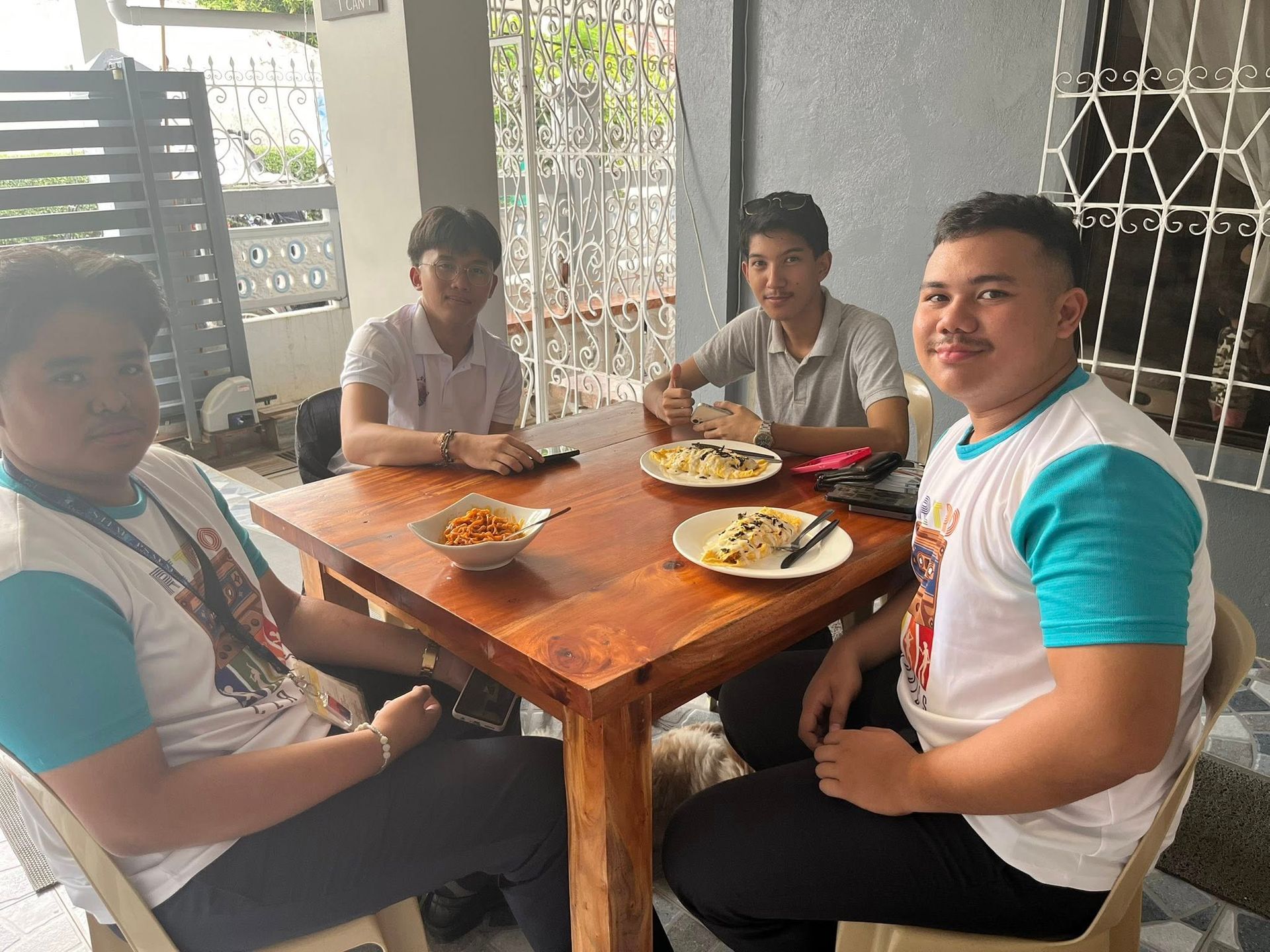Four people sit at a wooden square table eating meals on a covered patio with metal grill decor in the background.