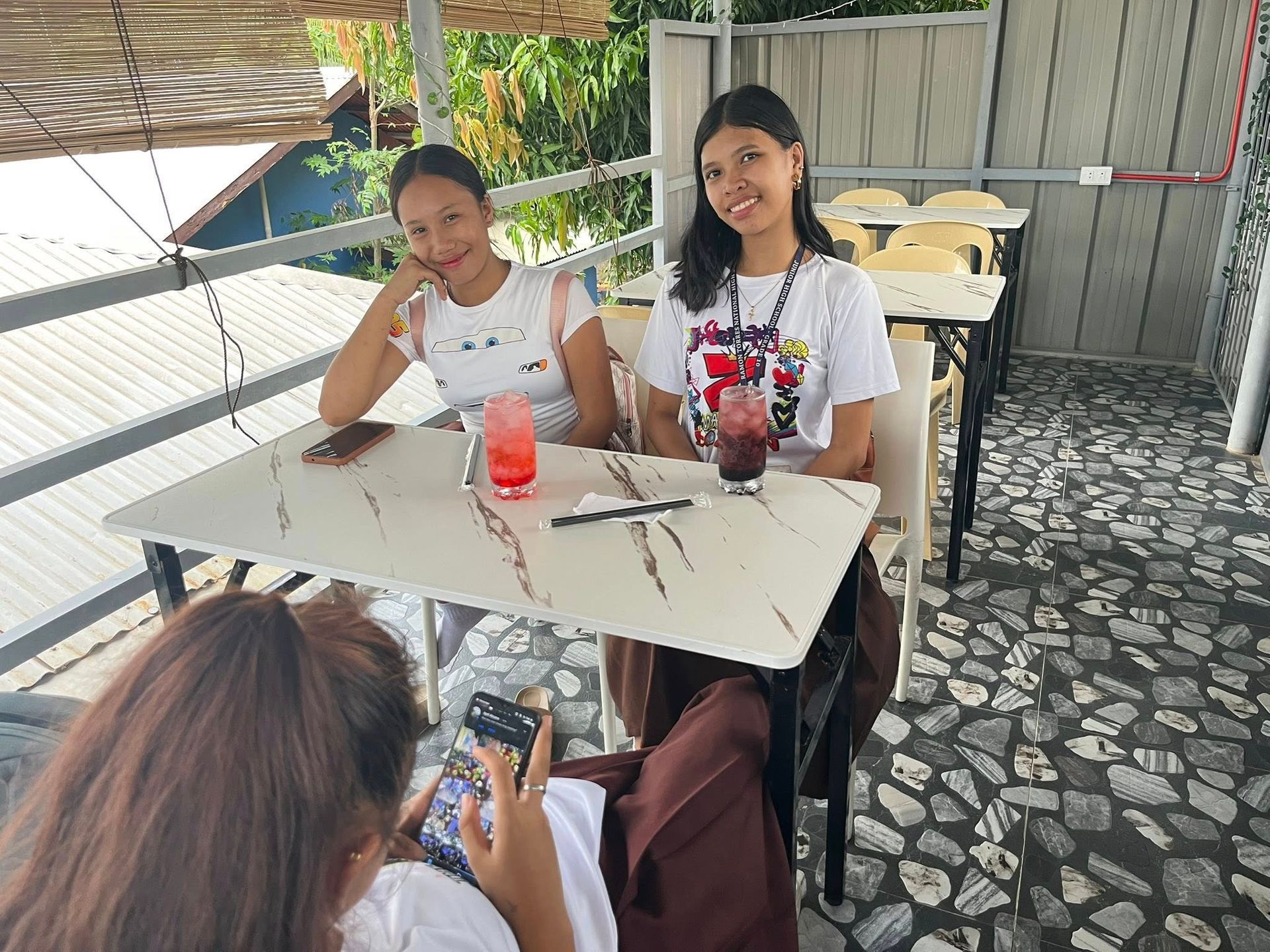 Three people sit at tables on a covered patio, with two facing the camera and one viewed from behind holding a phone.