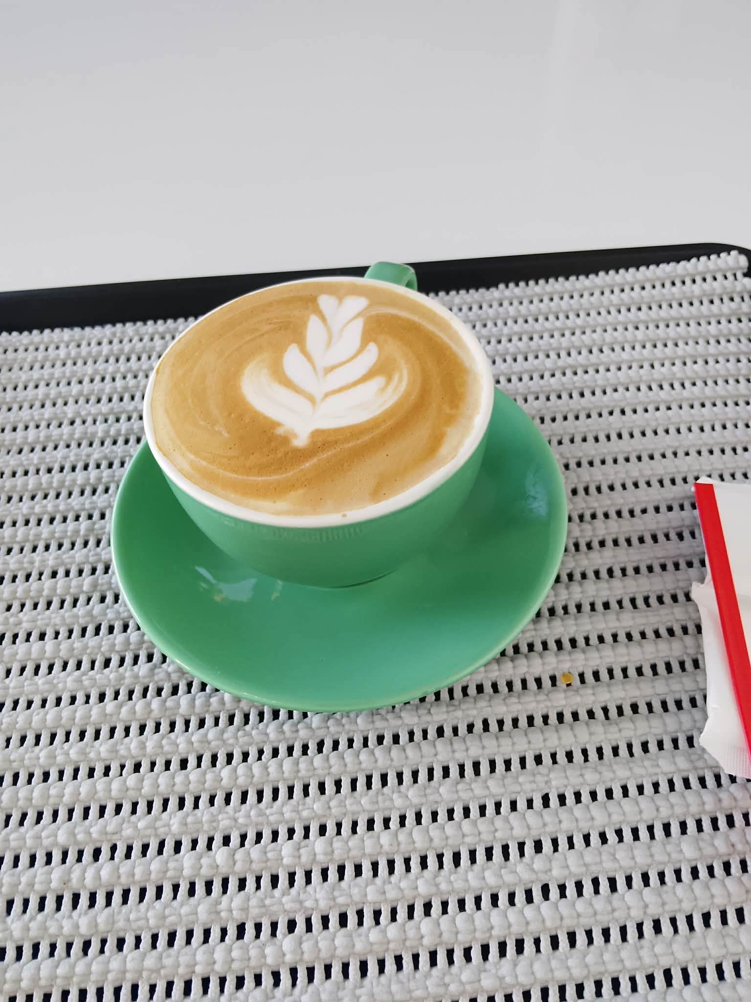 A light green cup and saucer containing a latte with white leaf-patterned foam art, resting on a textured gray placemat.