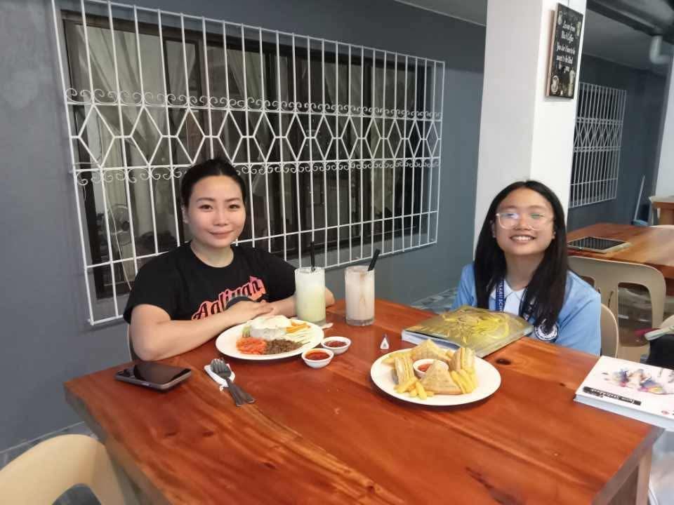 Two people sit at a wooden table in a cafe, smiling behind plates of food and glasses of drinks.