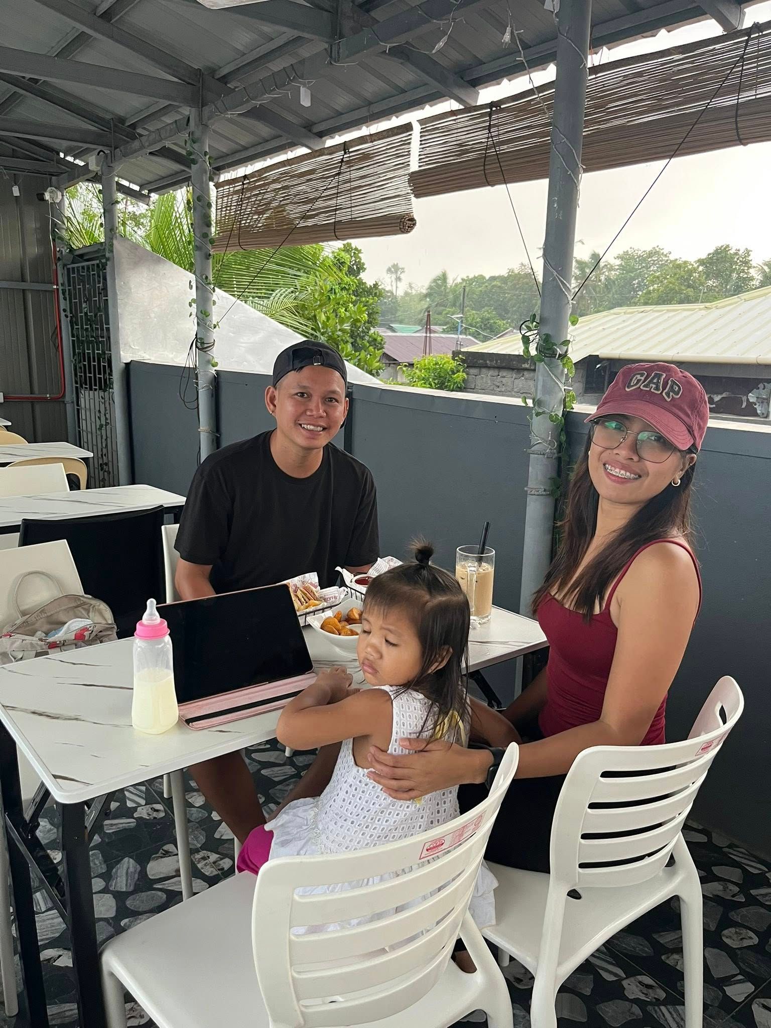 A family of three sitting at a table on a covered outdoor patio, with food, drinks, and a tablet on the table.