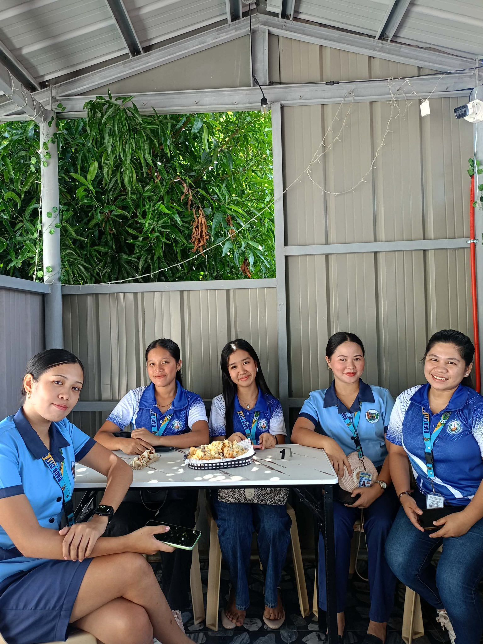 Five people in blue polo uniforms sit together at a table under an outdoor shelter, some smiling at the camera.