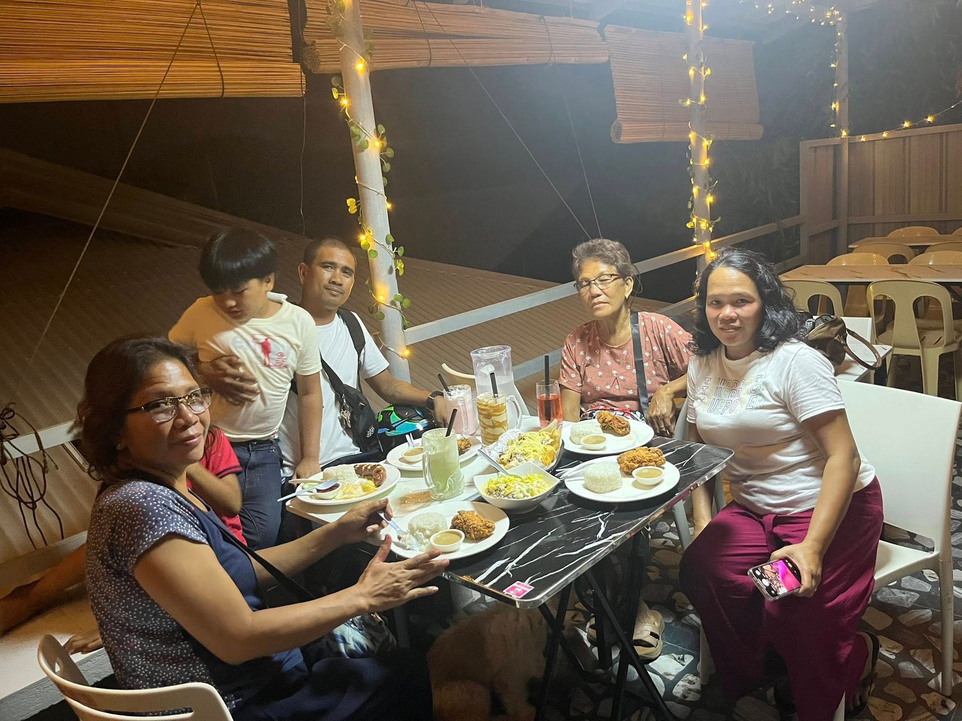 A family dining at an outdoor table with string lights, sitting on chairs and eating together at night.