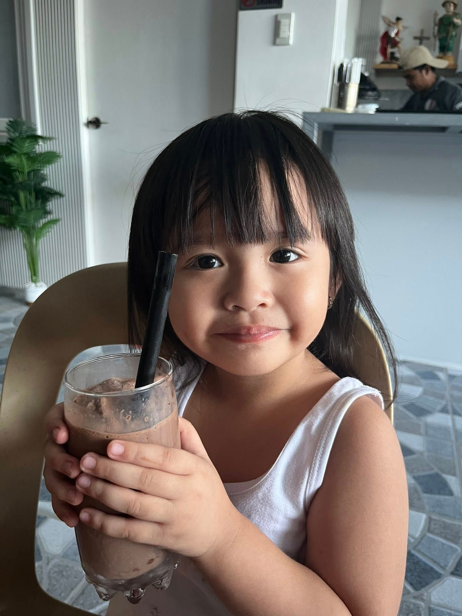 A child with dark hair smiles while holding a glass of chocolate drink with a straw in a bright, indoor setting.