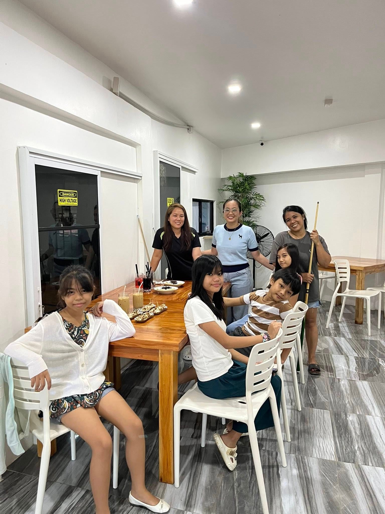 A group of people gathers around a wooden table in a bright, modern room, with some holding pool cues.