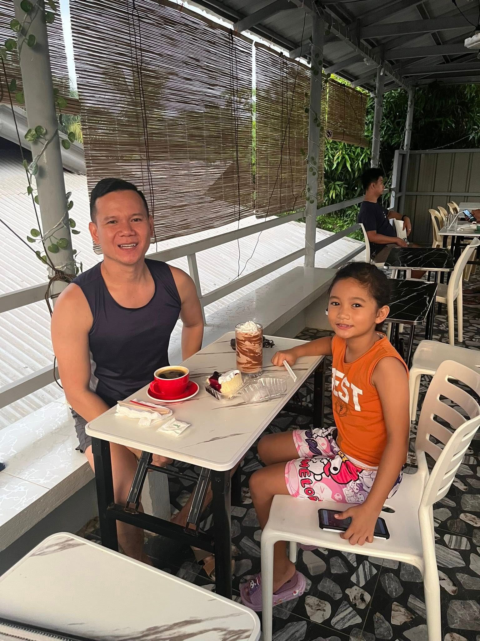 A man and a child sitting at a table with drinks in an open-air cafe, looking toward the camera.