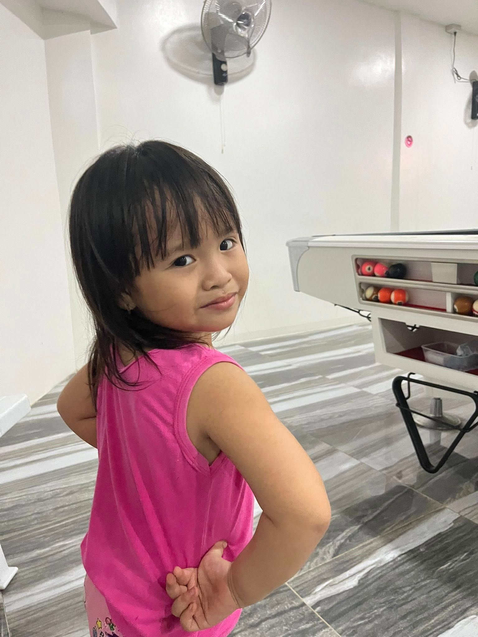 A young person with dark hair wearing a bright pink tank top looks over their shoulder near a pool table.