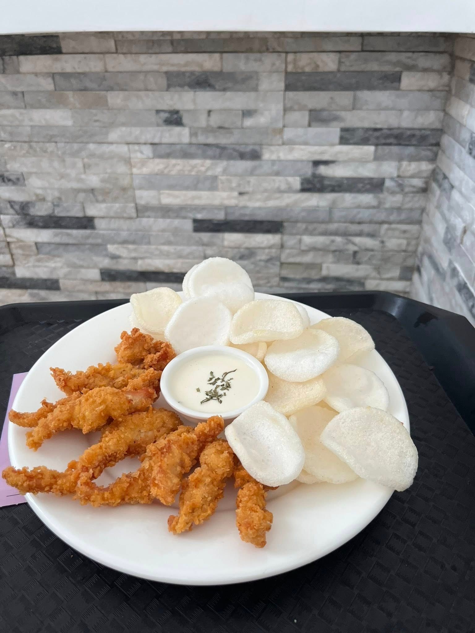 A white plate of crispy breaded chicken tenders, a small dish of creamy dipping sauce, and side of white rice crackers.