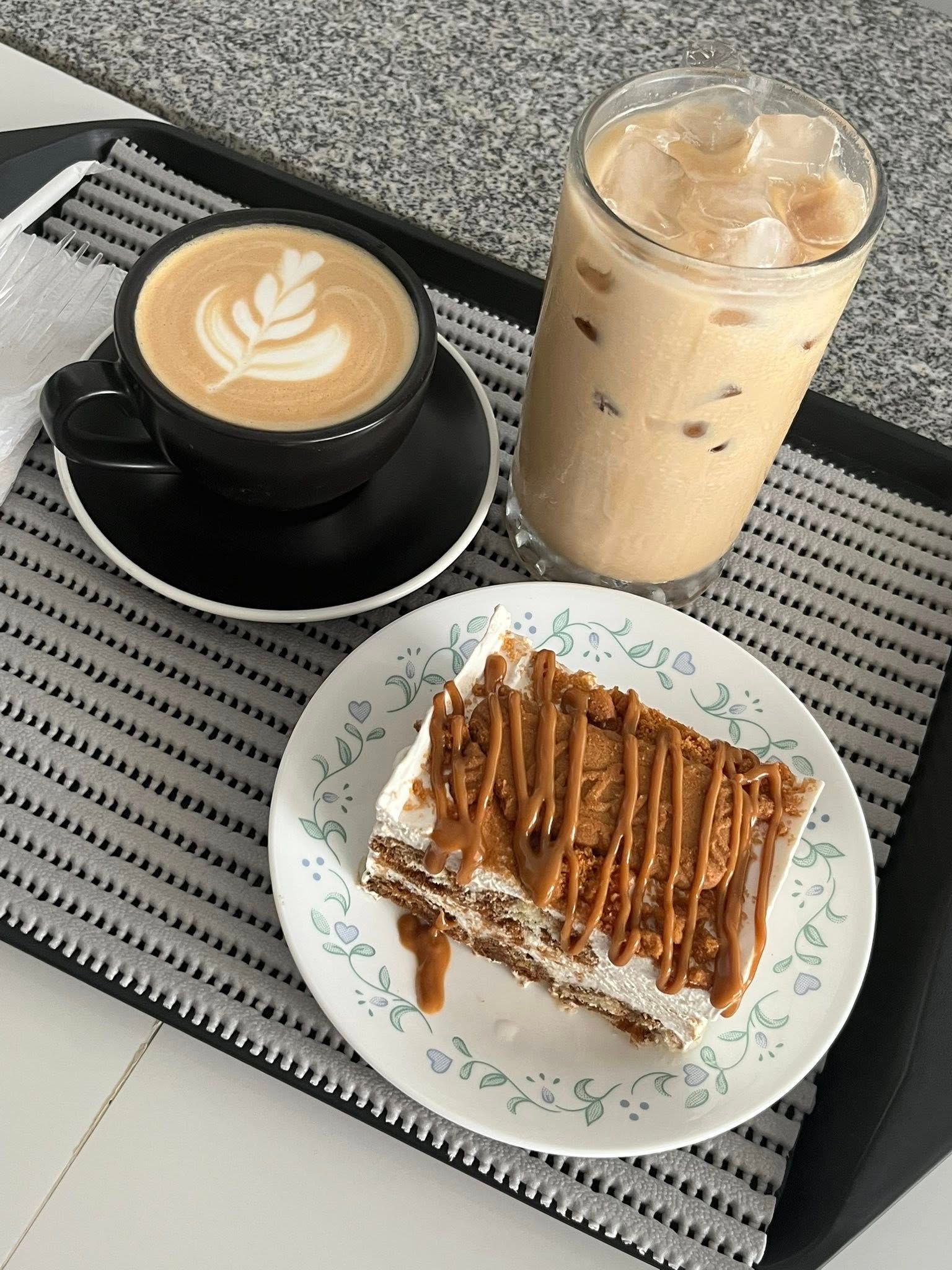 A tray with a hot latte with latte art, an iced coffee in a tall glass, and a square slice of caramel cake on a plate.