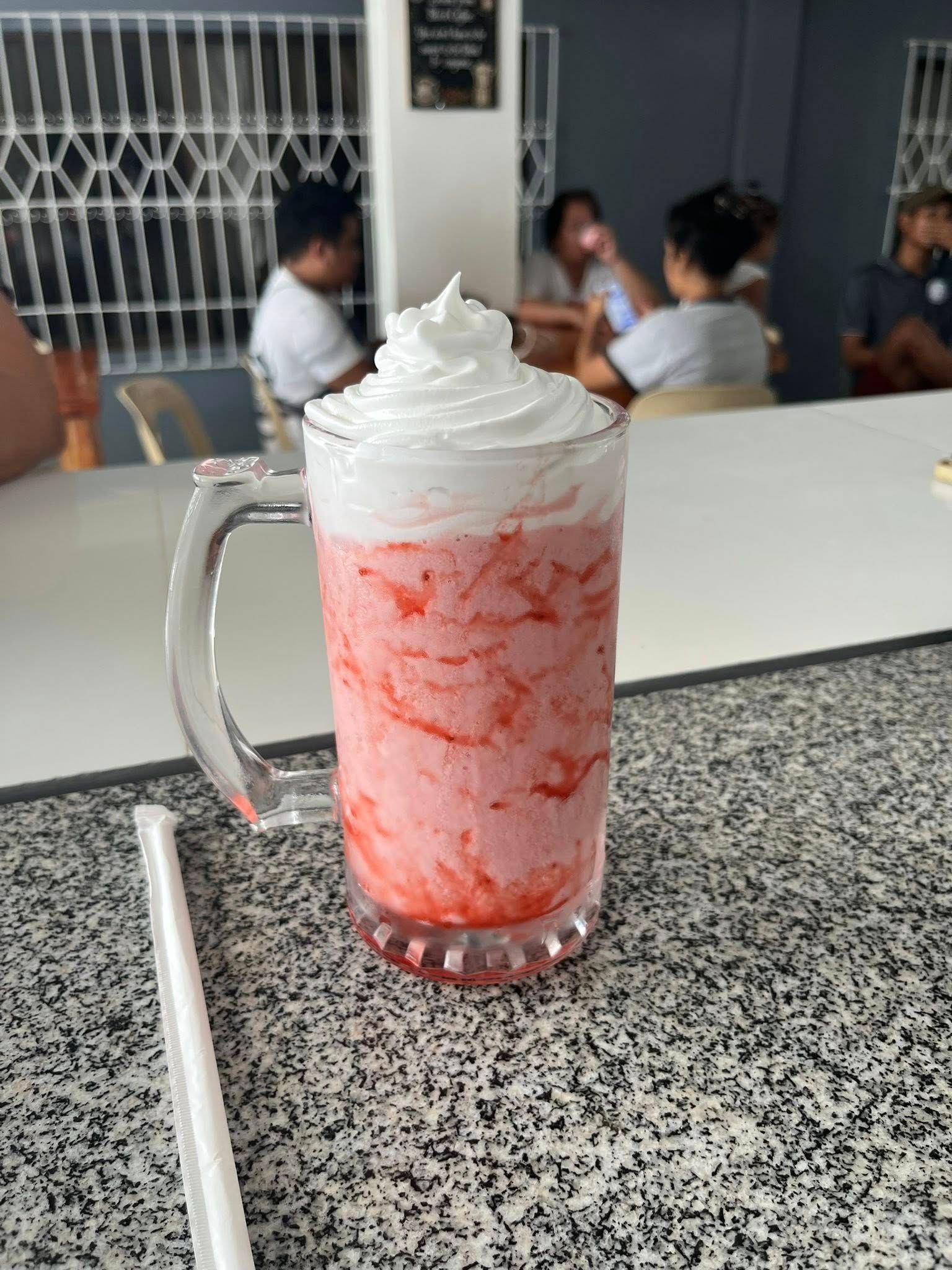 A frosted glass mug of pink strawberry smoothie topped with whipped cream, sitting on a speckled counter in a cafe.