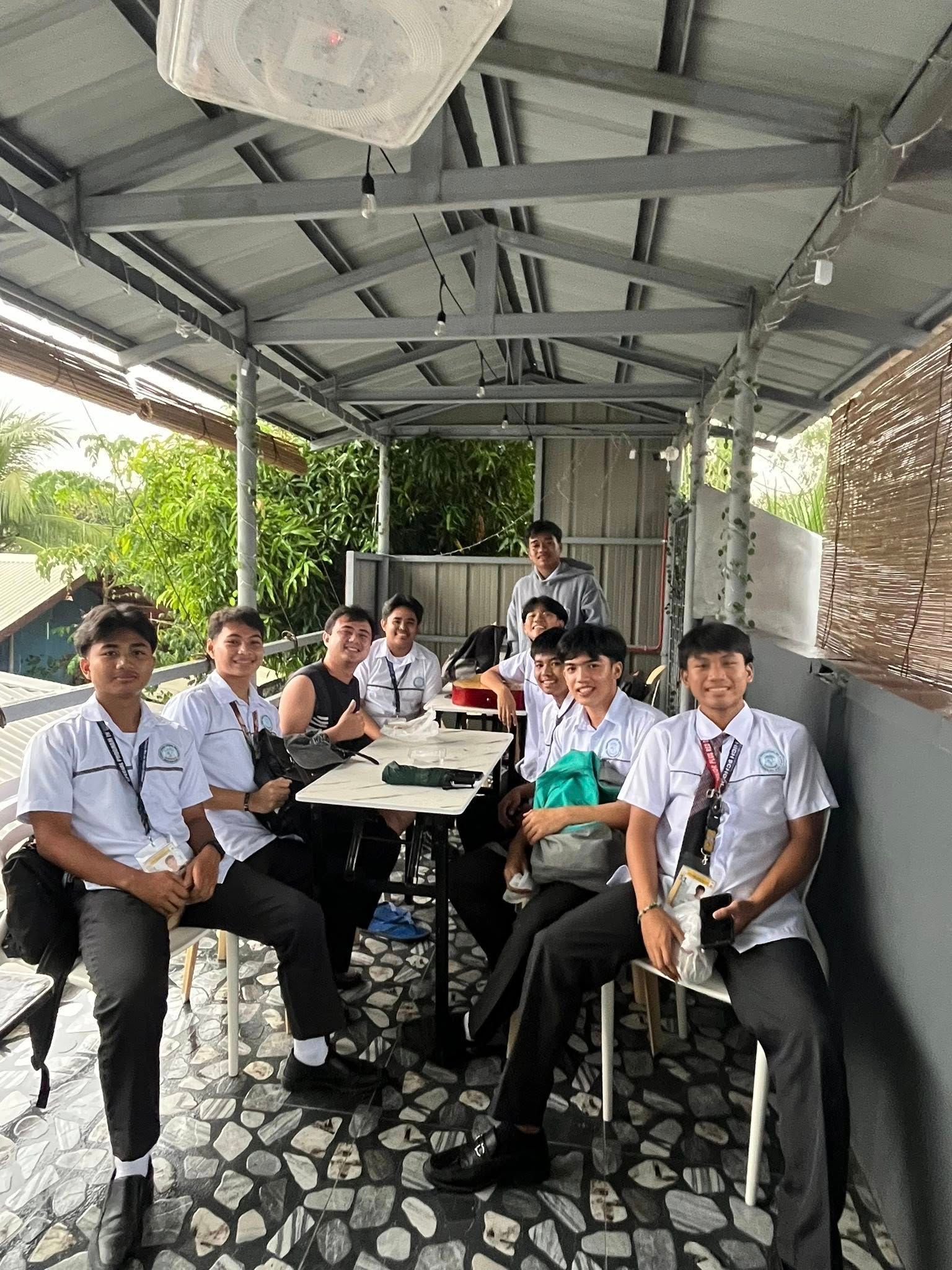 Students in white school uniforms sit at tables under an open-air pavilion.