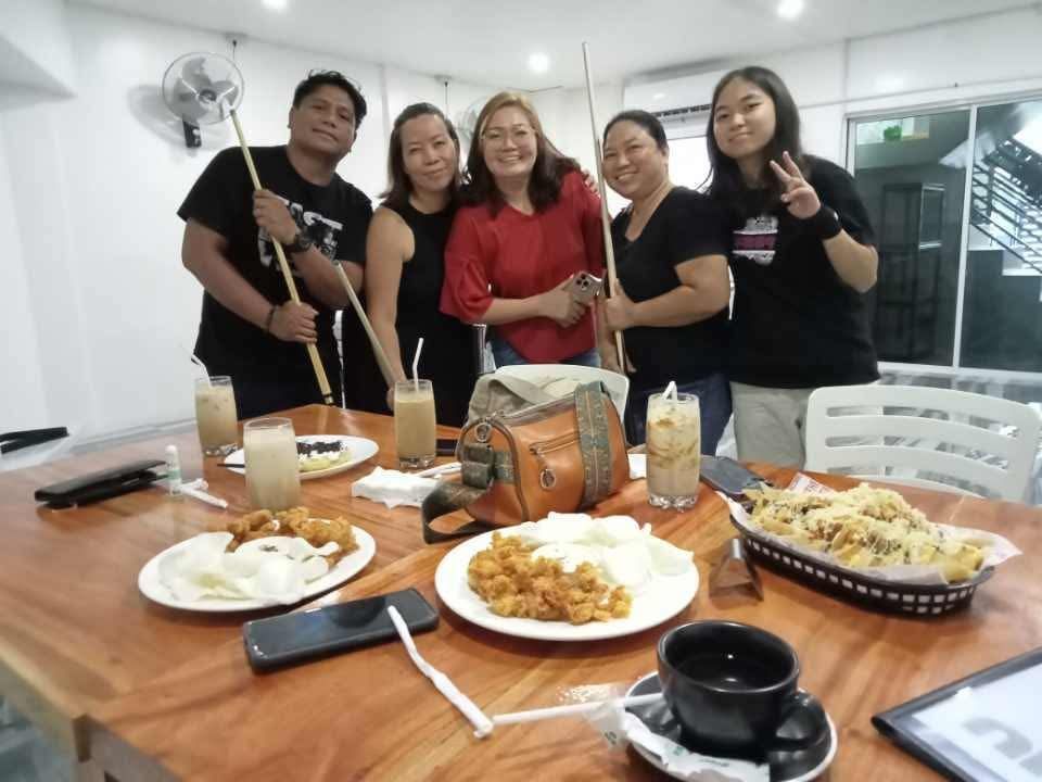 Five people stand smiling behind a wooden table topped with plates of food and drinks in a brightly lit indoor space.