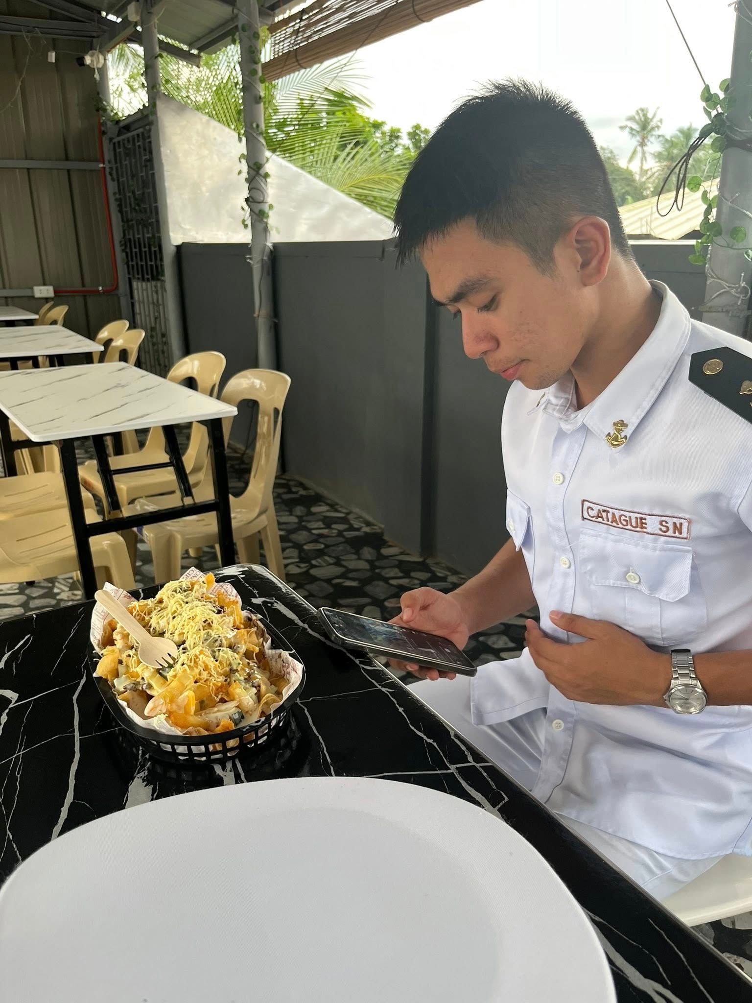 A person in a white uniform sits at a table, looking down at their phone next to a bowl of dessert in an outdoor cafe.
