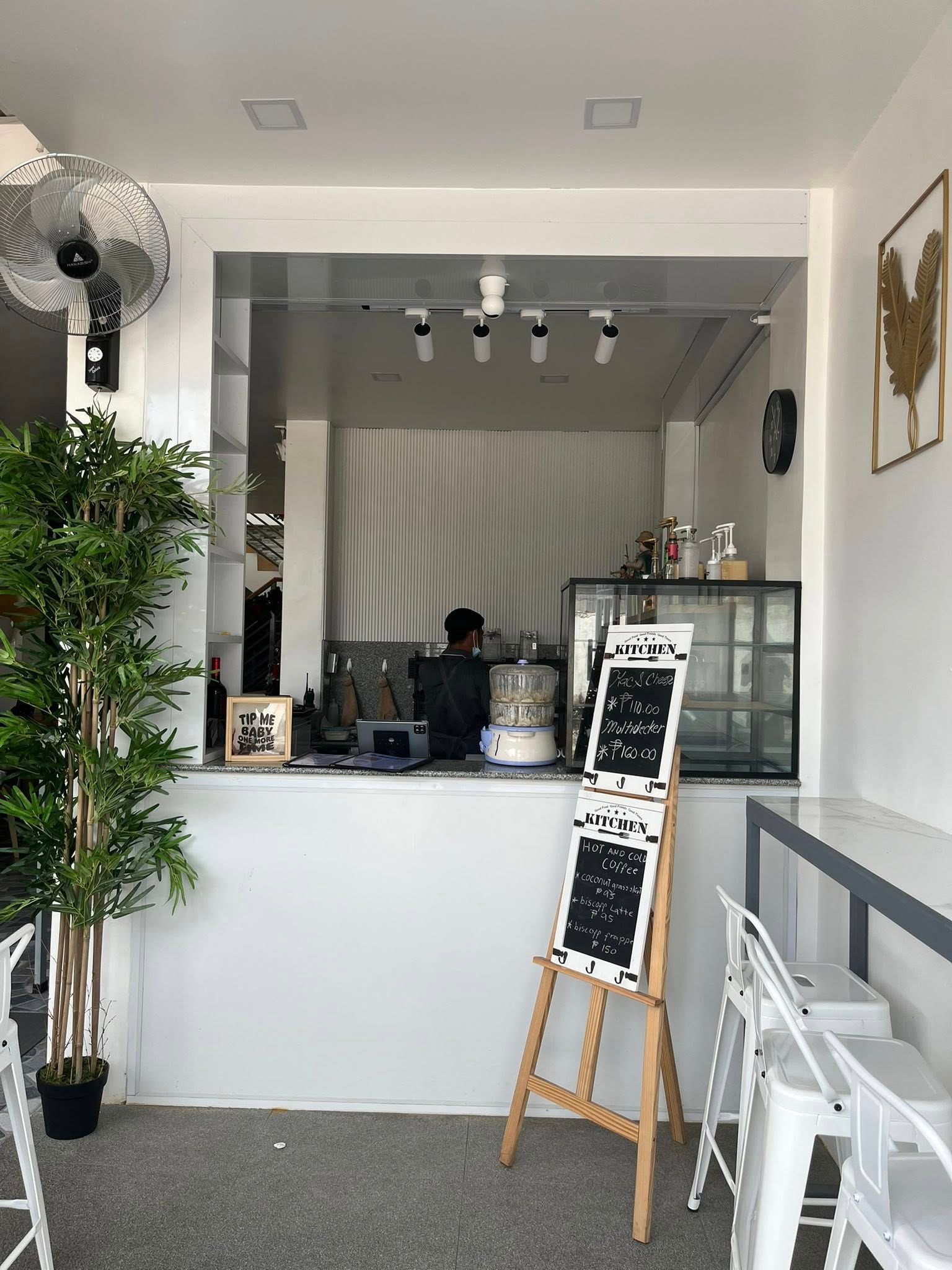 A counter in a brightly lit, modern cafe with a barista working behind it, a menu on an easel, and white seating.