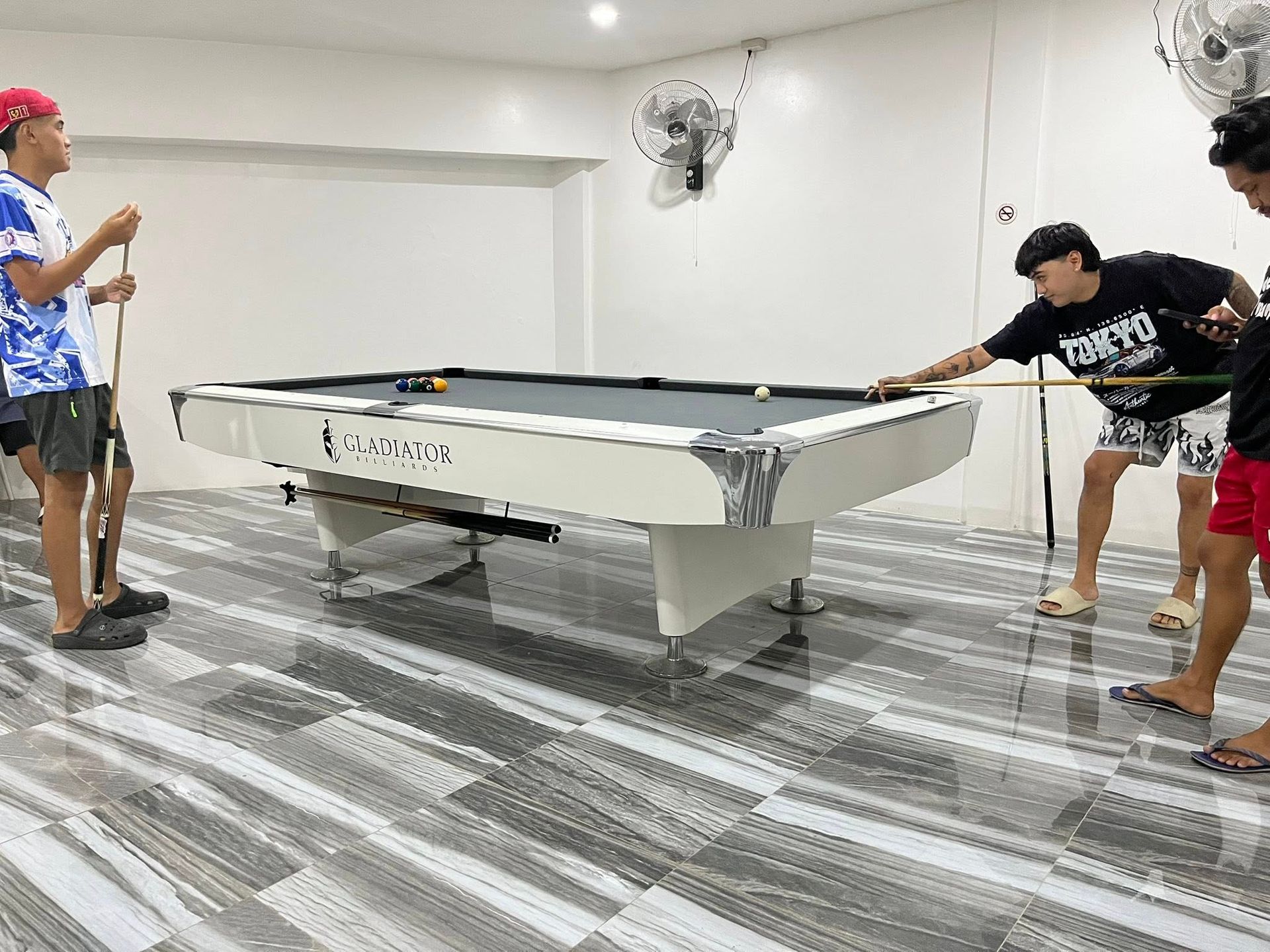 Three people playing billiards on a white table in a room with light-colored tile floors and two wall-mounted fans.