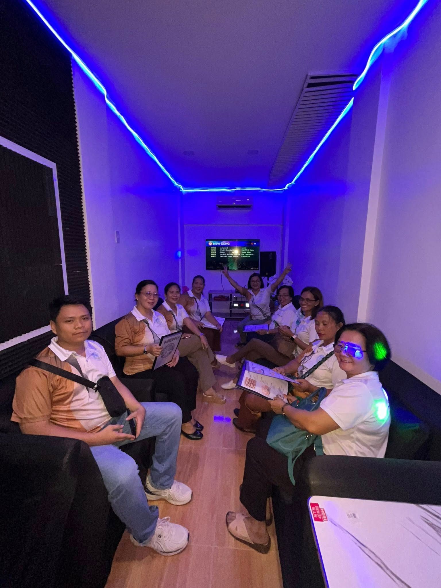 A group of people sit on couches inside a narrow, dimly lit room illuminated by blue LED strip lights along the ceiling.