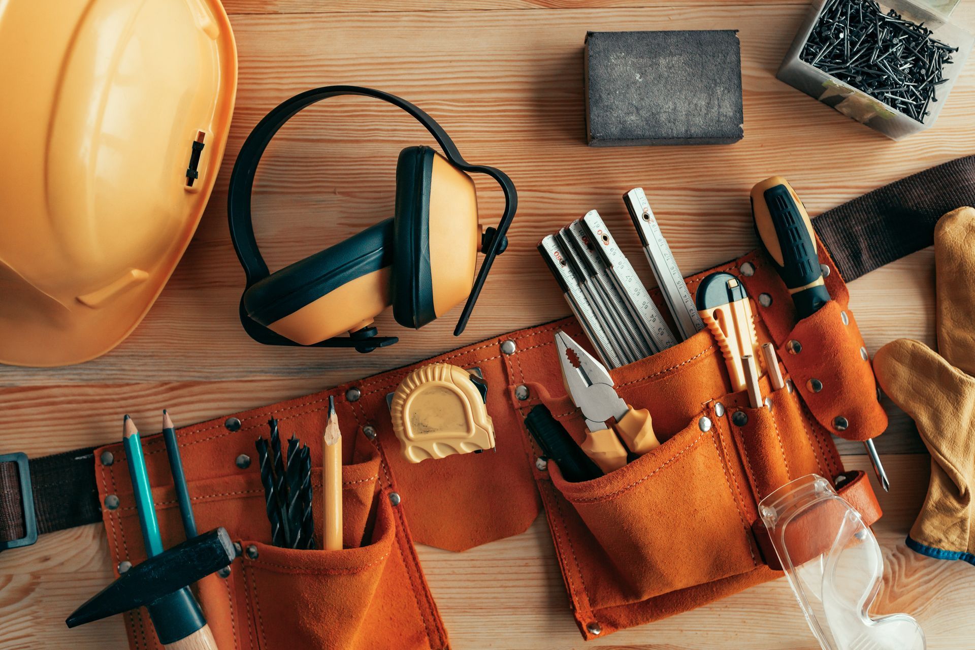 Work tools arranged on a wooden surface: hard hat, ear protection, tool belt with tools, sandpaper, nails, gloves.