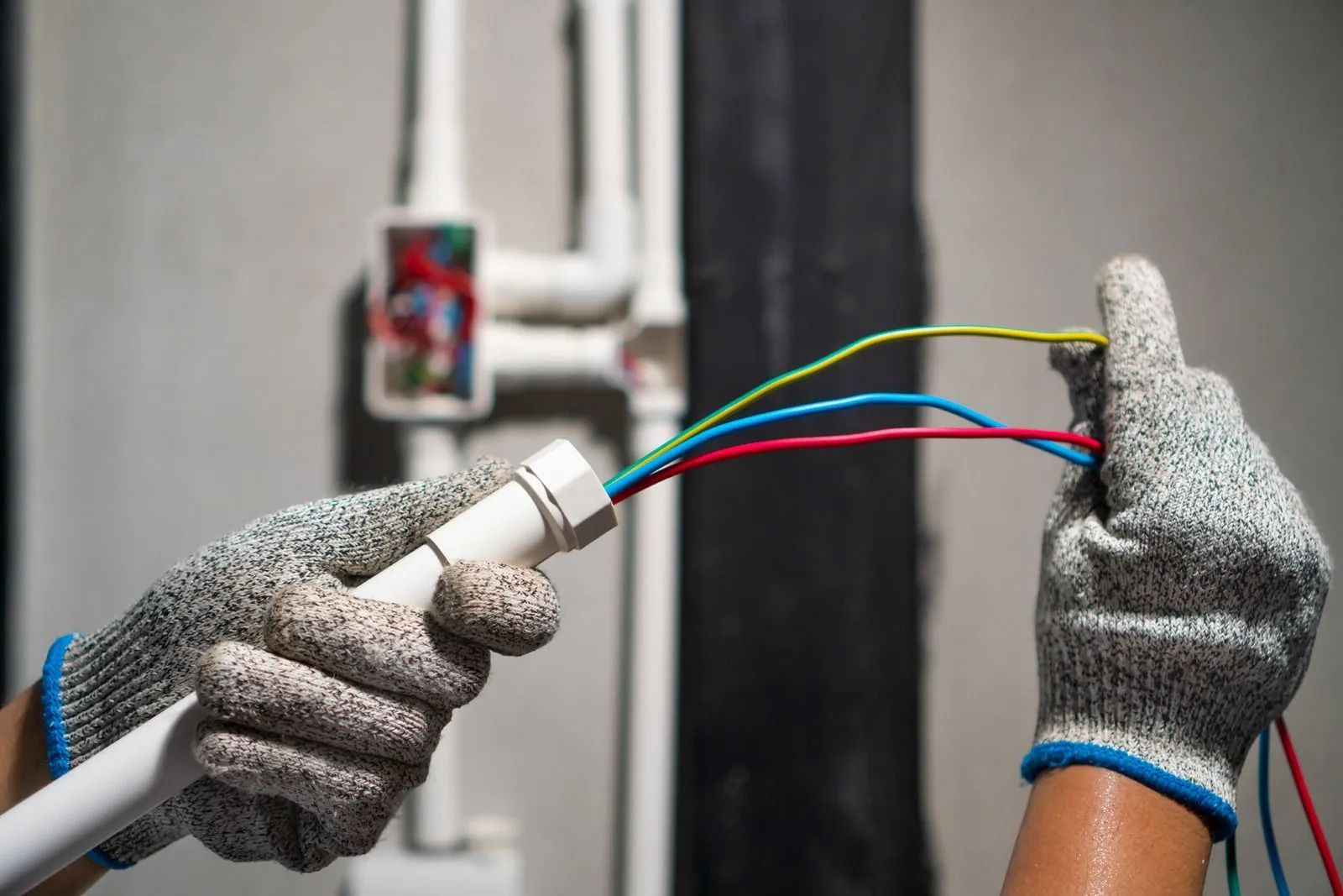 Hands in gray safety gloves pulling red, blue, and yellow electrical wires through a white conduit in a wall.
