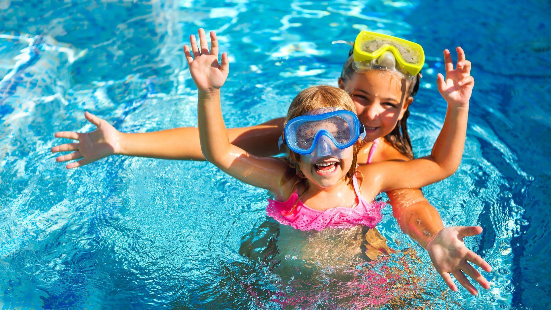 Two little girls wearing goggles are swimming in a pool.