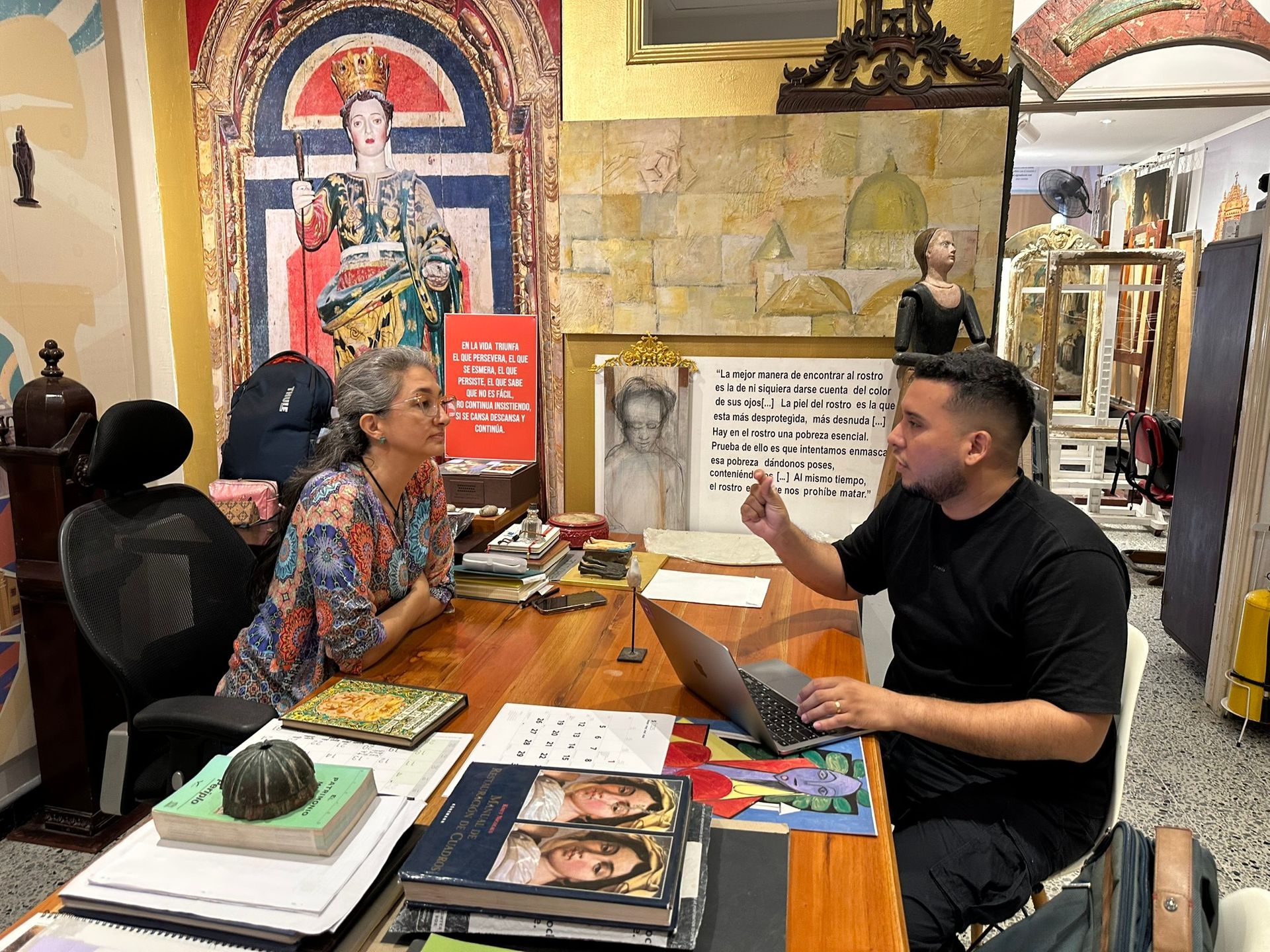 Mujer y hombre en un escritorio con una computadora portátil, discutiendo en una sala llena de arte.
