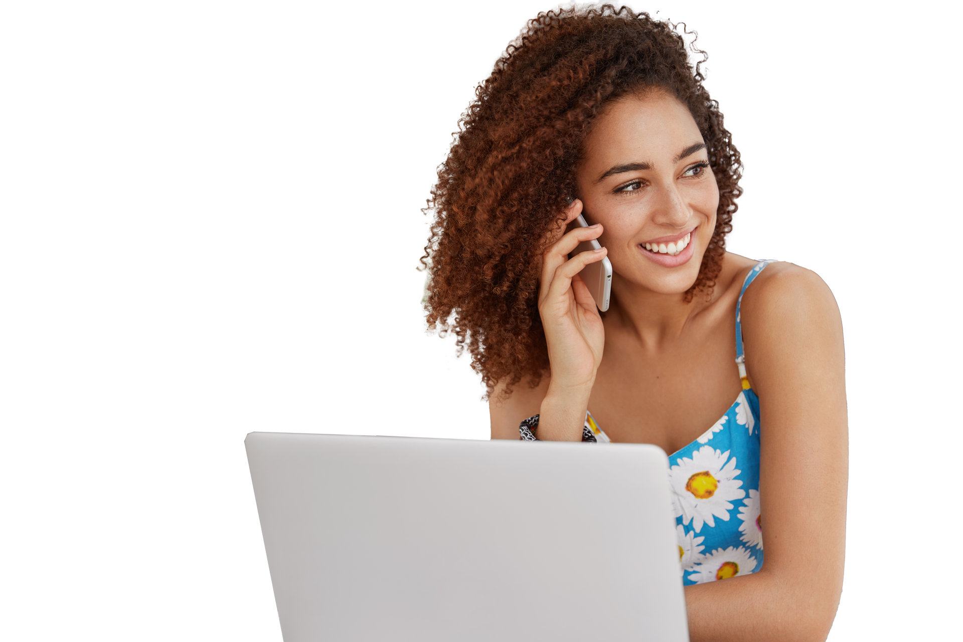 Mujer de cabello rizado, sonriendo, hablando por celular junto a una computadora portátil. 