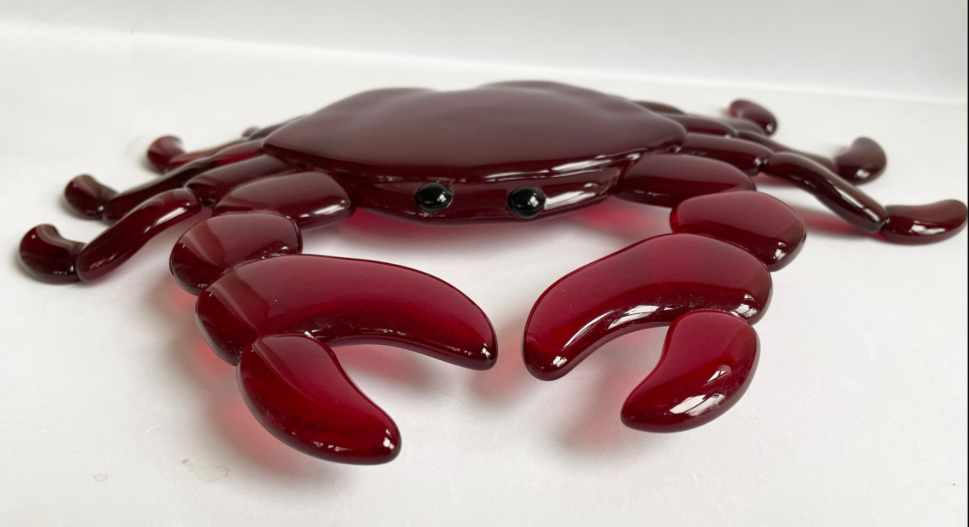 a red fused glass crab with black eyes sits on a white surface