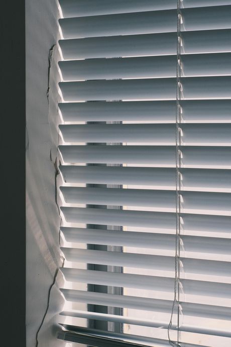 Horizontal white window blinds with a dark, jagged crack or mark running vertically along the adjacent wall.