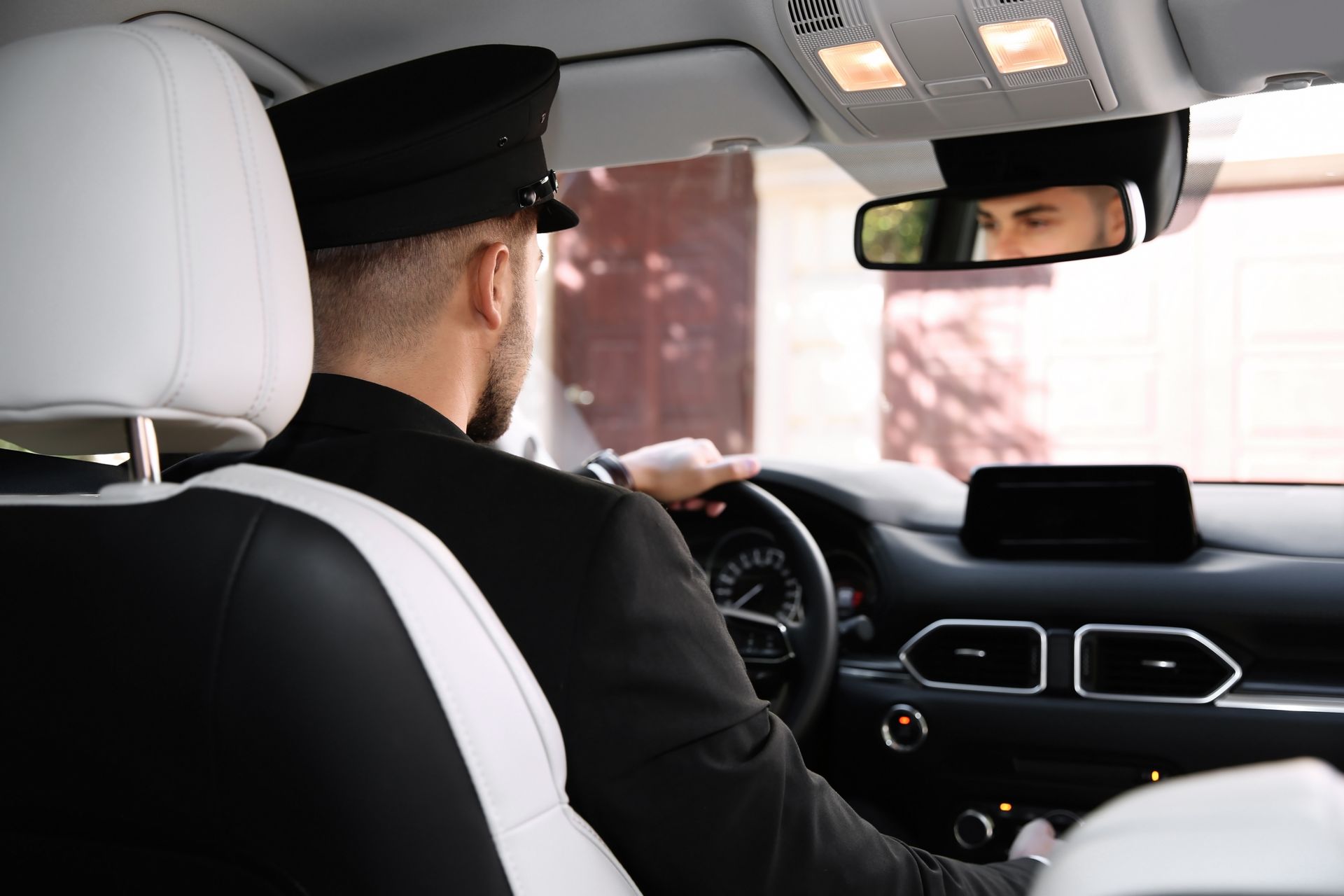 Man in uniform driving a car, seen from the back seat. He wears a hat and looks ahead, hands on the wheel.