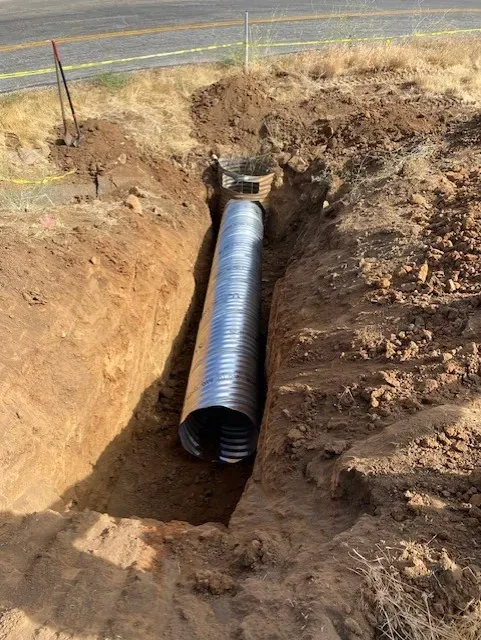 A corrugated metal pipe in an open trench alongside a road. Dirt and dry grass surround it.