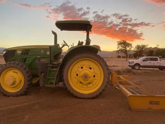 John Deere tractor with a yellow blade on a dirt lot at sunset. A white truck is parked nearby.