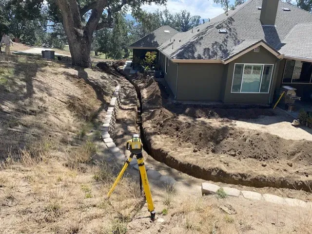 Trench dug beside a house, with a yellow surveying device on a tripod; sunny outdoor setting.
