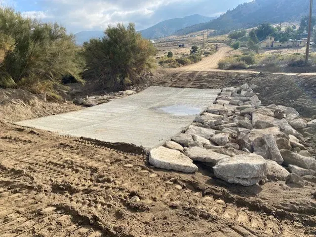 A concrete bridge with stone edging and dirt surroundings, leading to a dirt road in a valley.