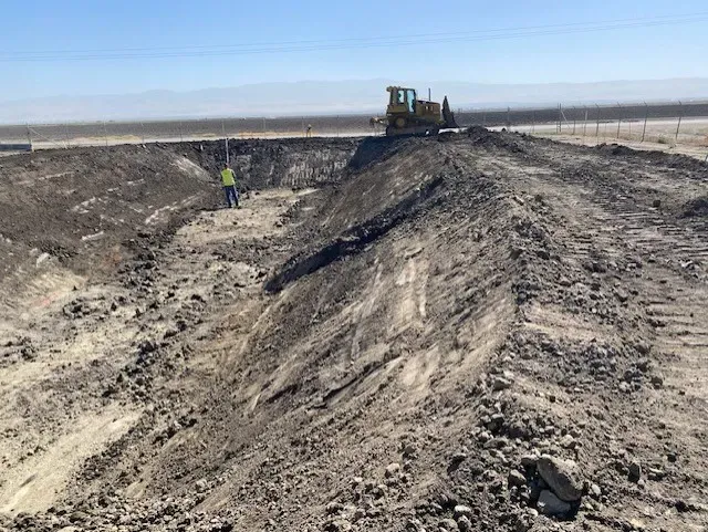 A bulldozer and worker excavate earth at a construction site in a field on a sunny day.
