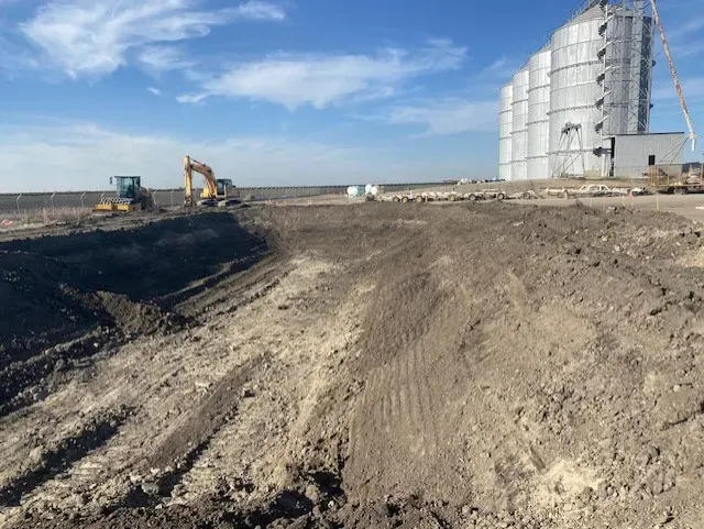 Construction site with dirt piles and excavators, grain silos in the background under a blue sky.