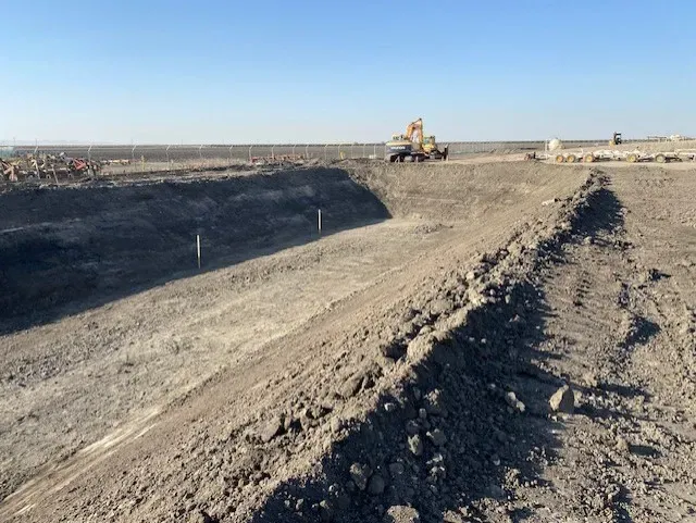Construction site: Excavated earth forming a wide trench with machinery in the background under a blue sky.