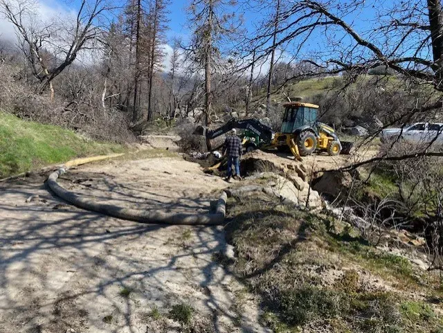 A person and backhoe repair a dirt road. A hose lies on the ground. Trees and hillside in the background.
