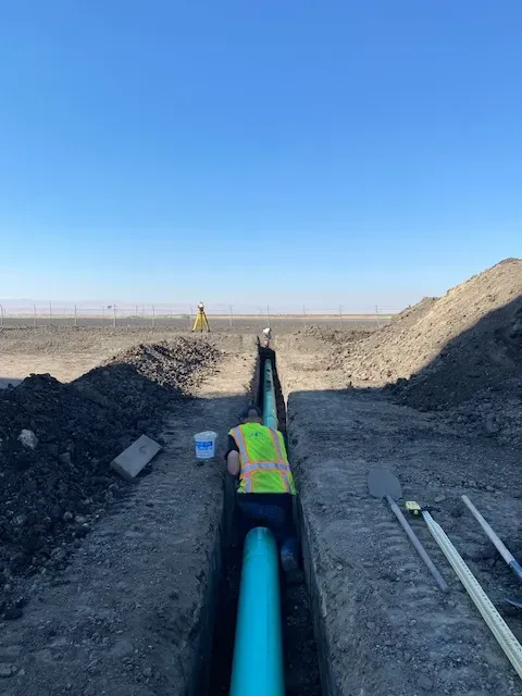 Construction worker in trench with blue pipe, another worker in background under blue sky.