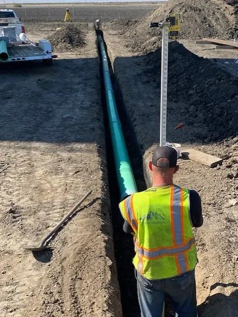 Construction worker in neon vest near a trench with a green pipe. Leveling stick and a truck present.