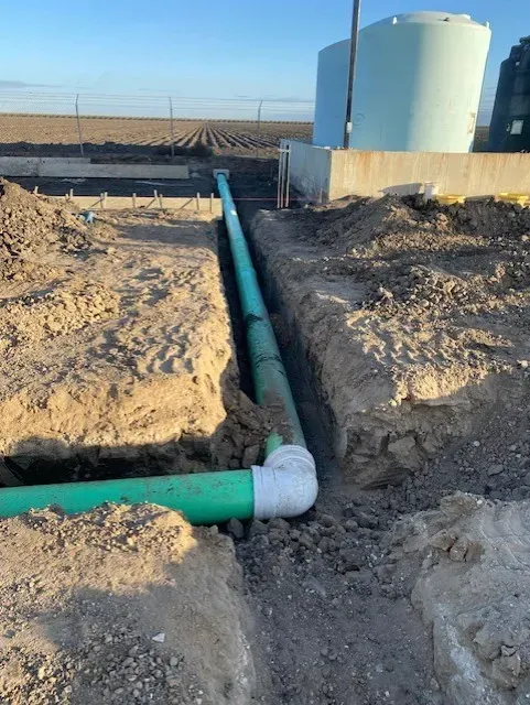 Green pipes in a trench, connected with a white elbow, near tanks and a field.
