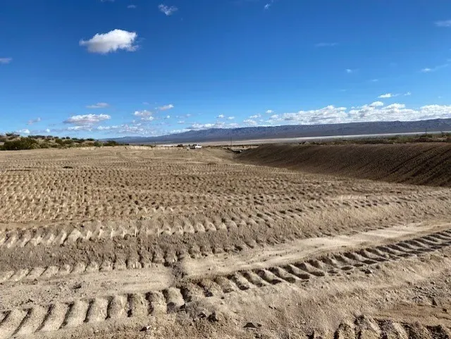 Tractor tracks on a cleared, graded field under a blue sky. Low hills and clouds in the distance.