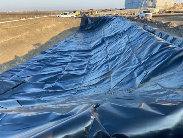 Black plastic liner being installed in a construction pit, with workers and vehicles visible in the background.