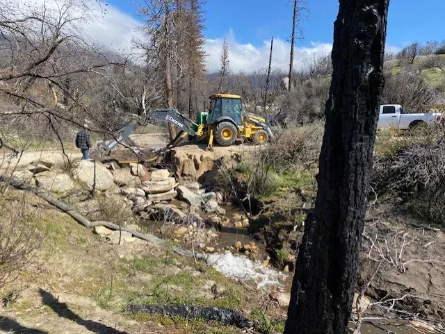 A backhoe repairs a road crossing a stream. Burned trees and a person stand nearby.