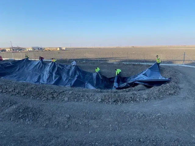 Workers in safety vests lay a large black tarp over a circular pit in a brown, barren landscape.