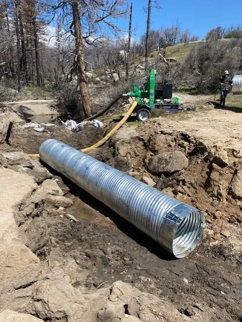 A corrugated metal culvert pipe is laid in a ditch with a pump nearby in a natural setting.