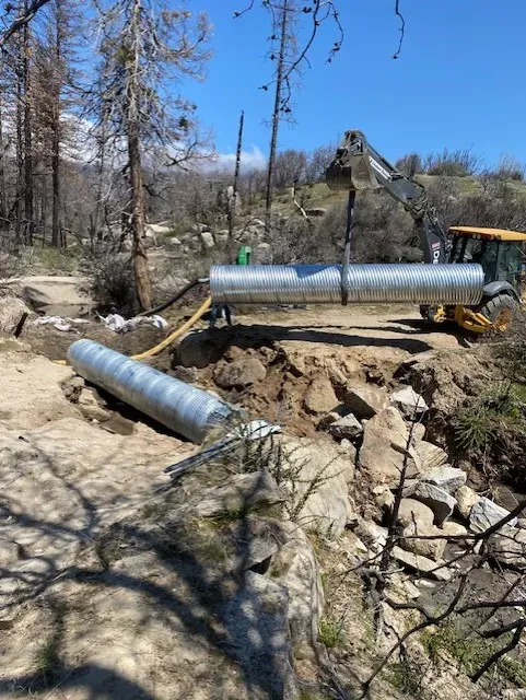 A tractor places a large metal culvert over a creek bed. Other culverts lie nearby. Sunny day.