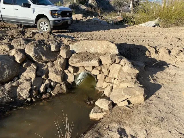 Rock-lined culvert under a dirt road, with a white pickup truck parked nearby.