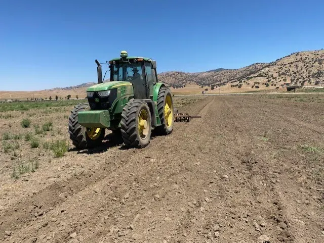 Green tractor plowing a brown field under a blue sky, with rolling hills in the background.