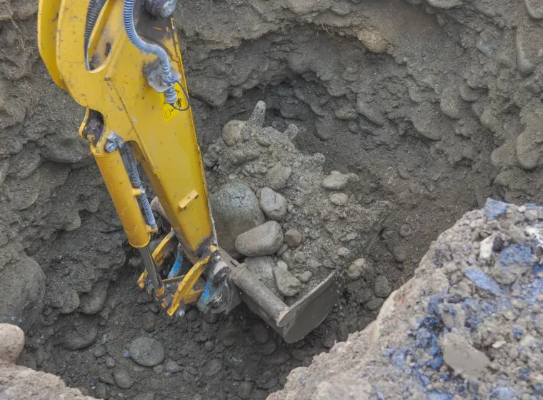 Yellow excavator bucket digging into a hole filled with soil and rocks.