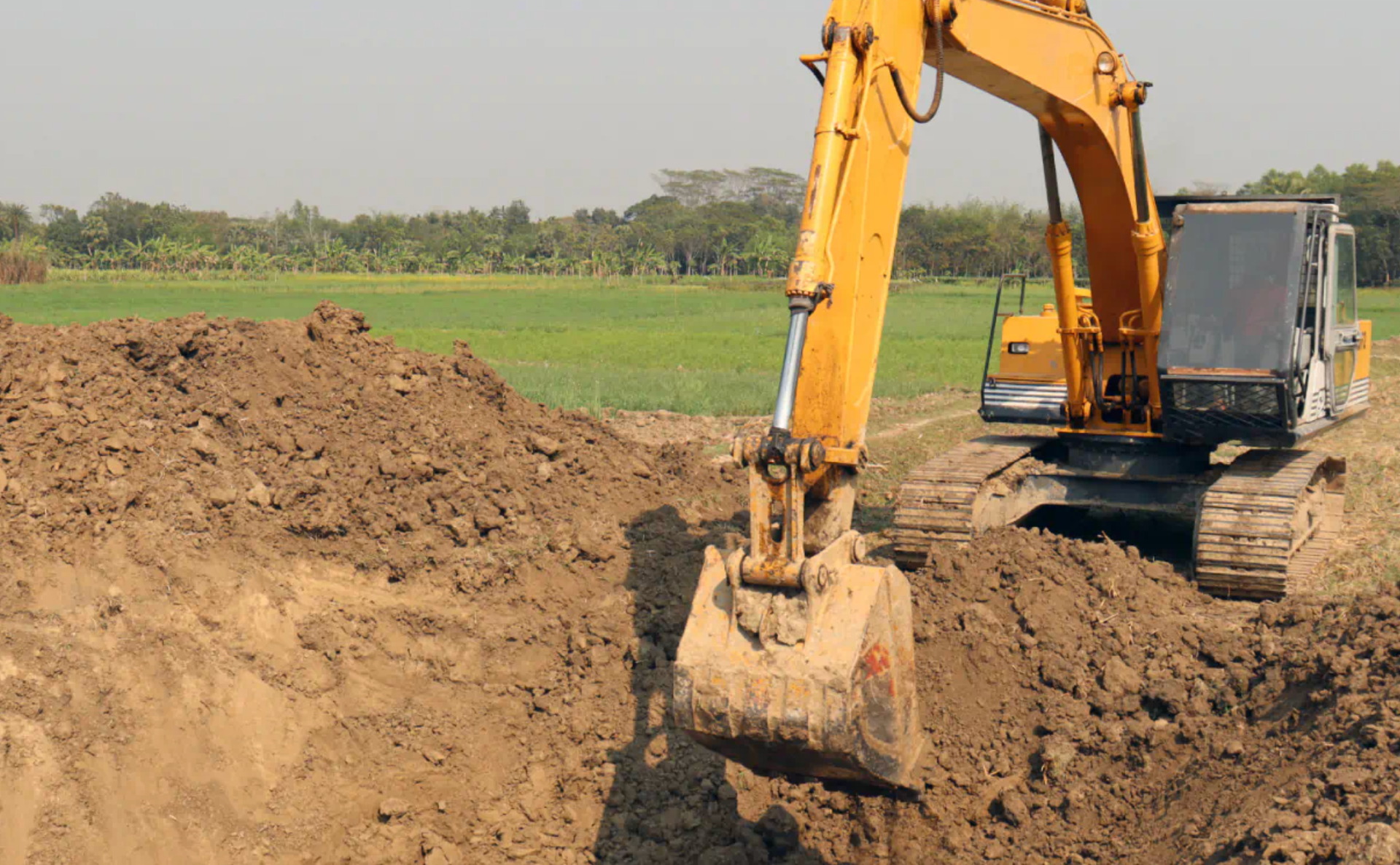 Yellow excavator digging a trench in a field. Brown soil, green grass, and trees are in the background.
